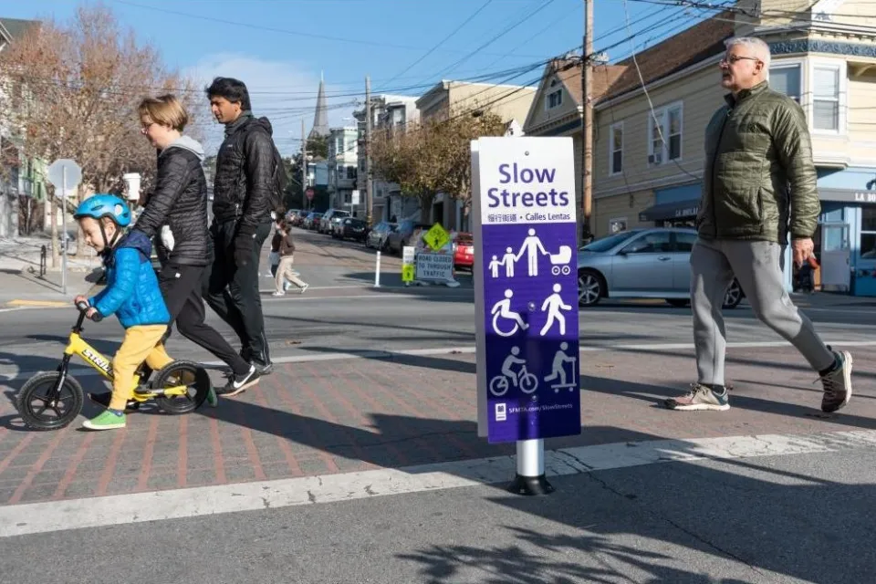 White and purple sign for Slow Street in San Francisco, California with people crossing crosswalk.